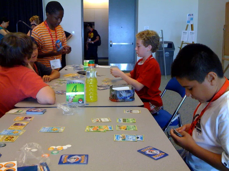 Two young players face off across a table covered in Pokemon Trading Card Game cards at a tournament.